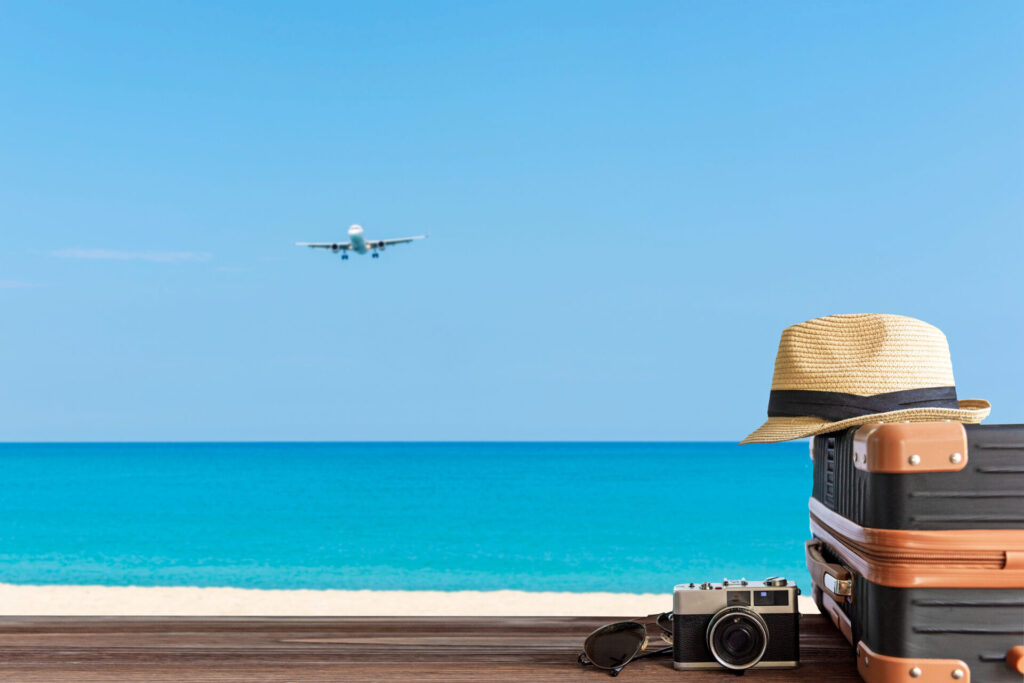 海辺に置かれたスーツケースと帽子、カメラ、空には飛行機
