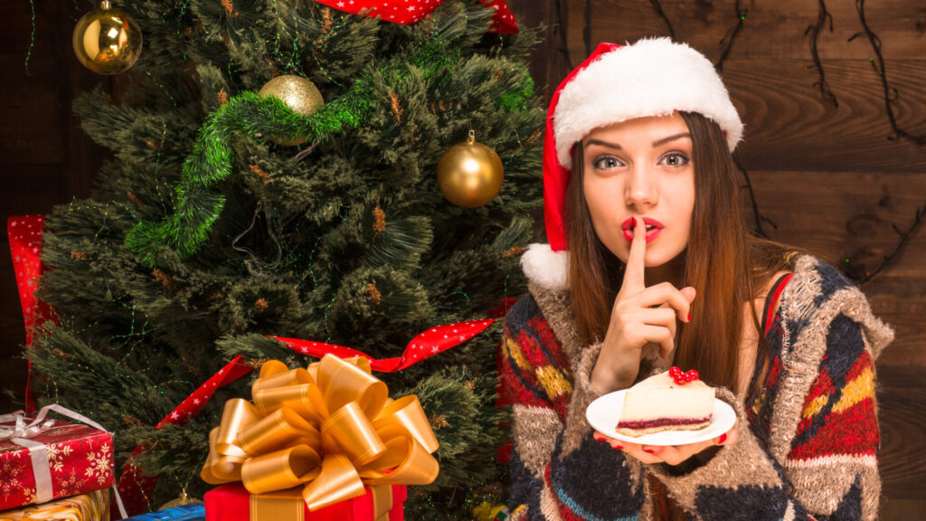 クリスマスツリー前でサンタ帽子をかぶりケーキを持ってシーっと人差し指を口元にあてる女性