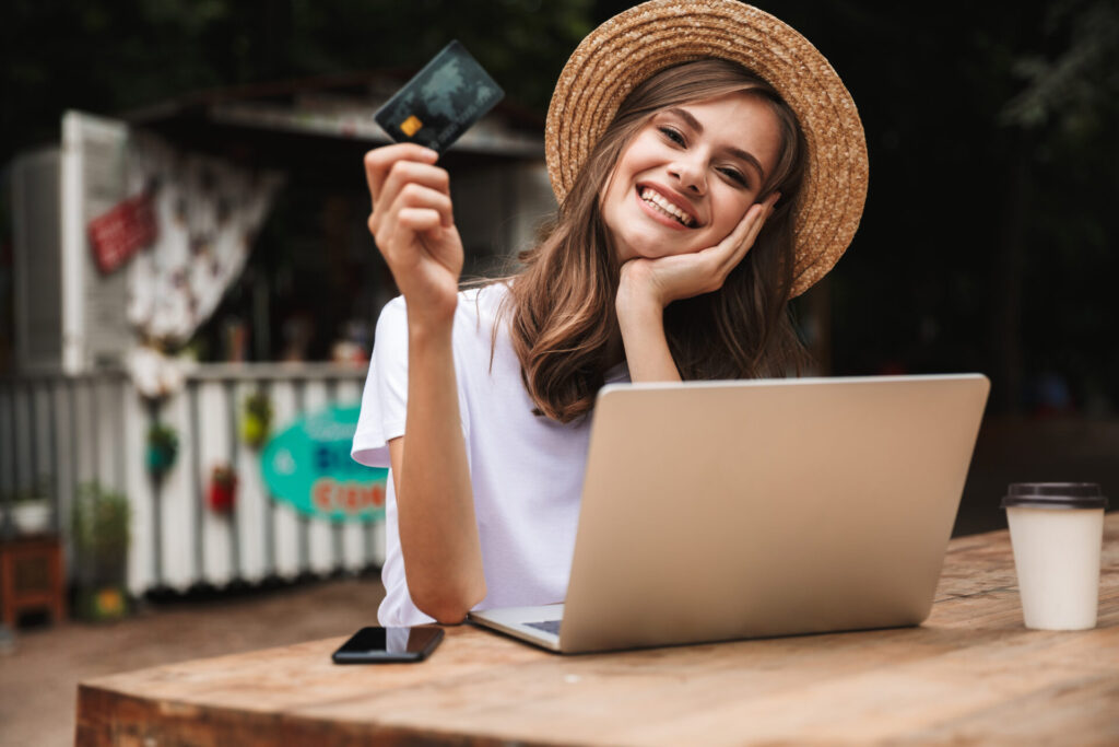 麦わら帽子をかぶり屋外のテーブルに肘をついてパソコン前に座るクレジットカードを持った微笑む女性