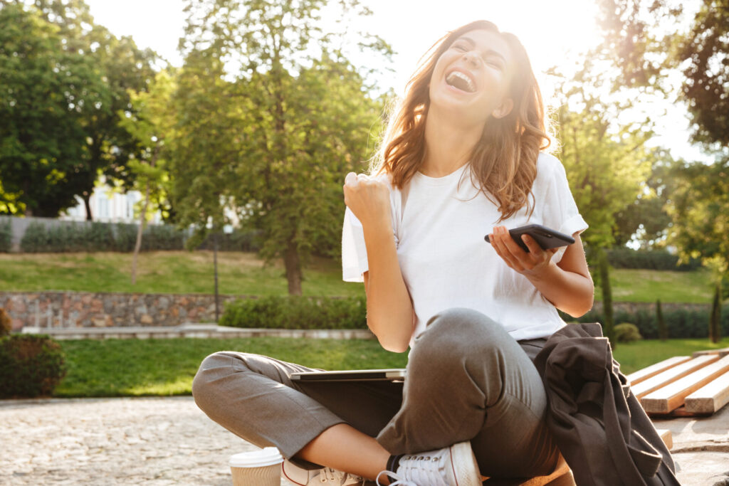 屋外でスマホを持ち座る女性がガッツポーズをしている